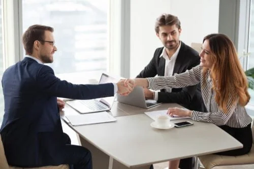A couple meeting with their attorney at a desk with laptops and documents. The woman and the attorney are shaking hands.