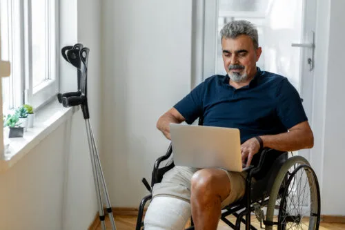 A man in a wheelchair with bandages on one leg, crutches next to him, and a computer in his lap.