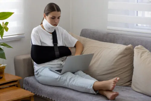 A woman reclining on a couch using the computer. Her arm is in a sling and her neck is in a brace.