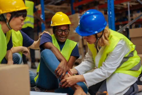 An injured worker holding their leg while being examined by a medical professional. 2 other workers are sitting nearby.