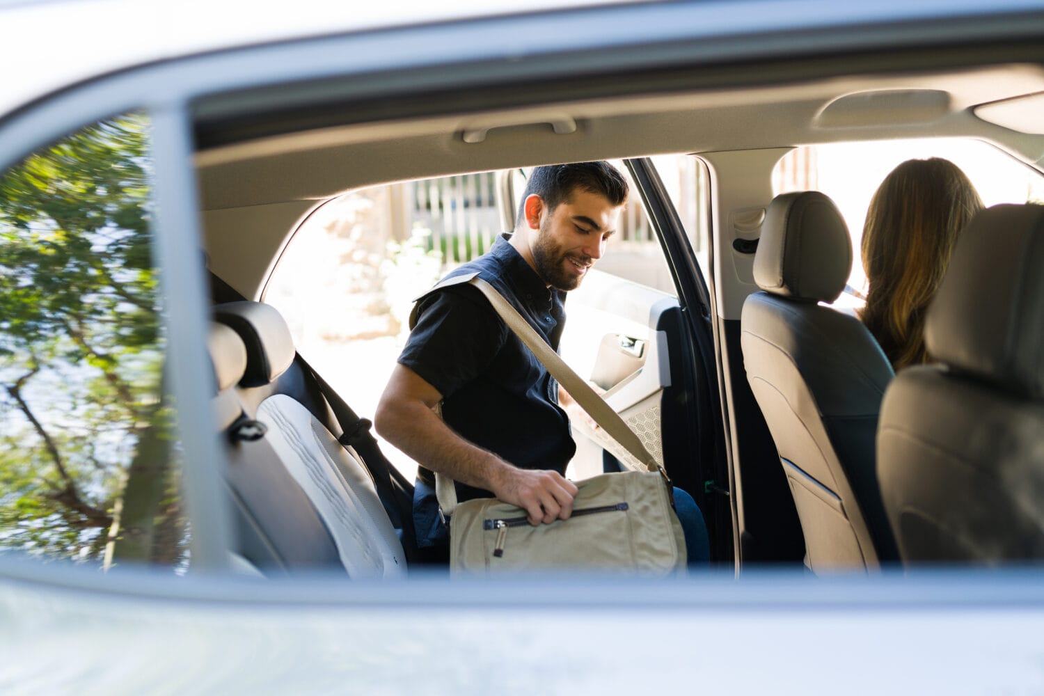A smiling man with a bag over his shoulder enters the backseat of a rideshare vehicle.