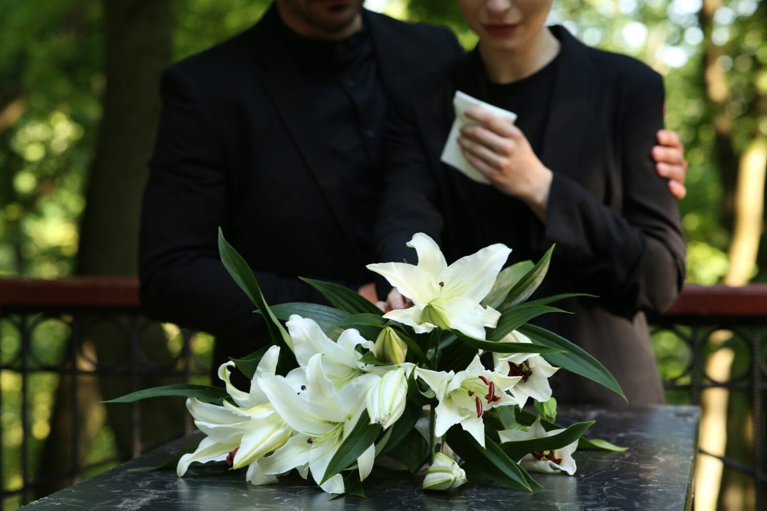 A couple in black clothes stand next to a casket topped with white flowers. The woman is holding a tissue.