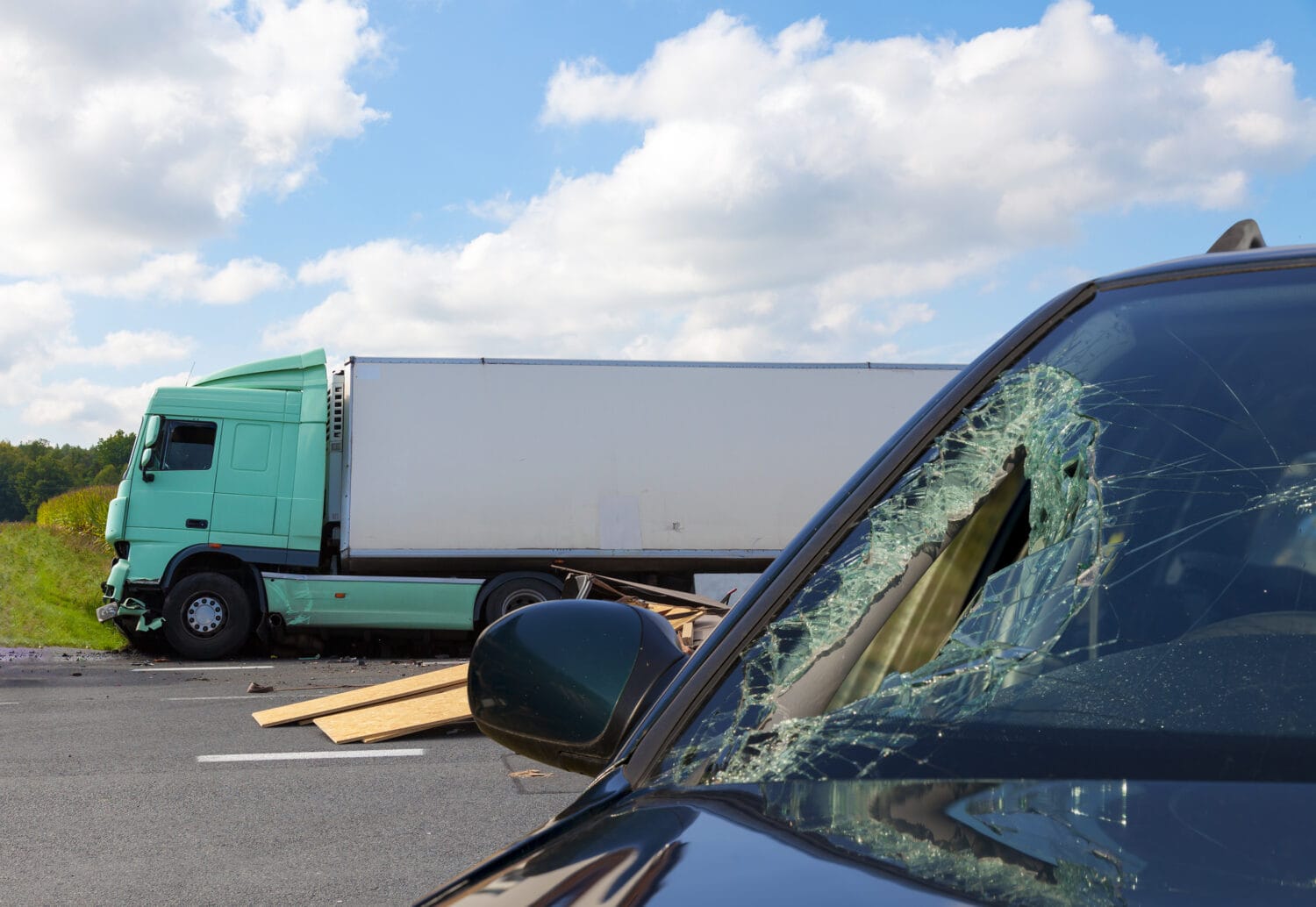 Close-up of a car’s damaged windshield, and a semi-truck on the side of a highway behind the car after a serious accident.