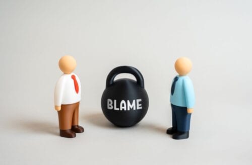 Toy people stand next to kettlebell labeled “Blame” as they argue over fault.