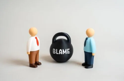 Toy people stand next to kettlebell labeled “Blame” as they argue over fault.