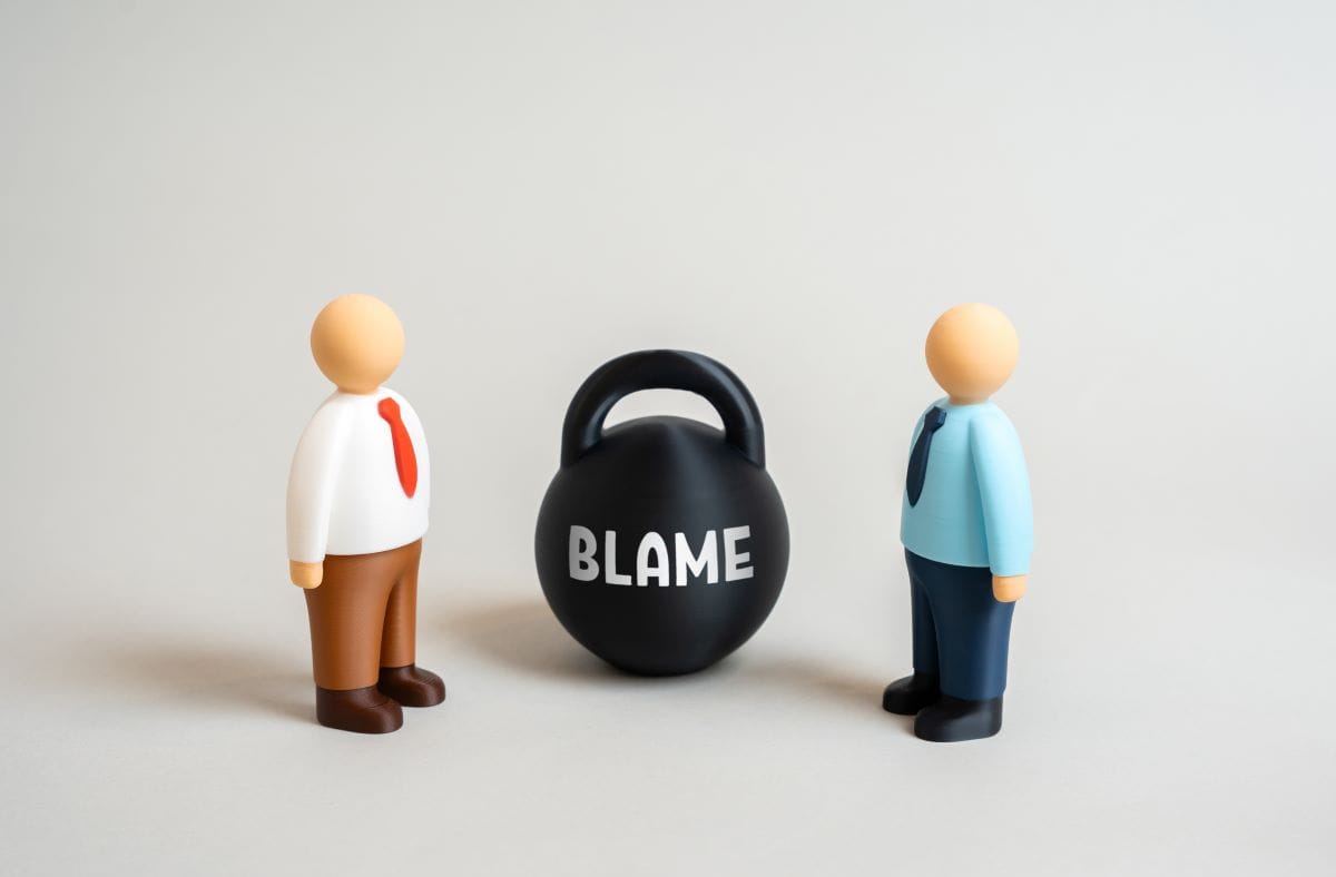 Toy people stand next to kettlebell labeled “Blame” as they argue over fault.