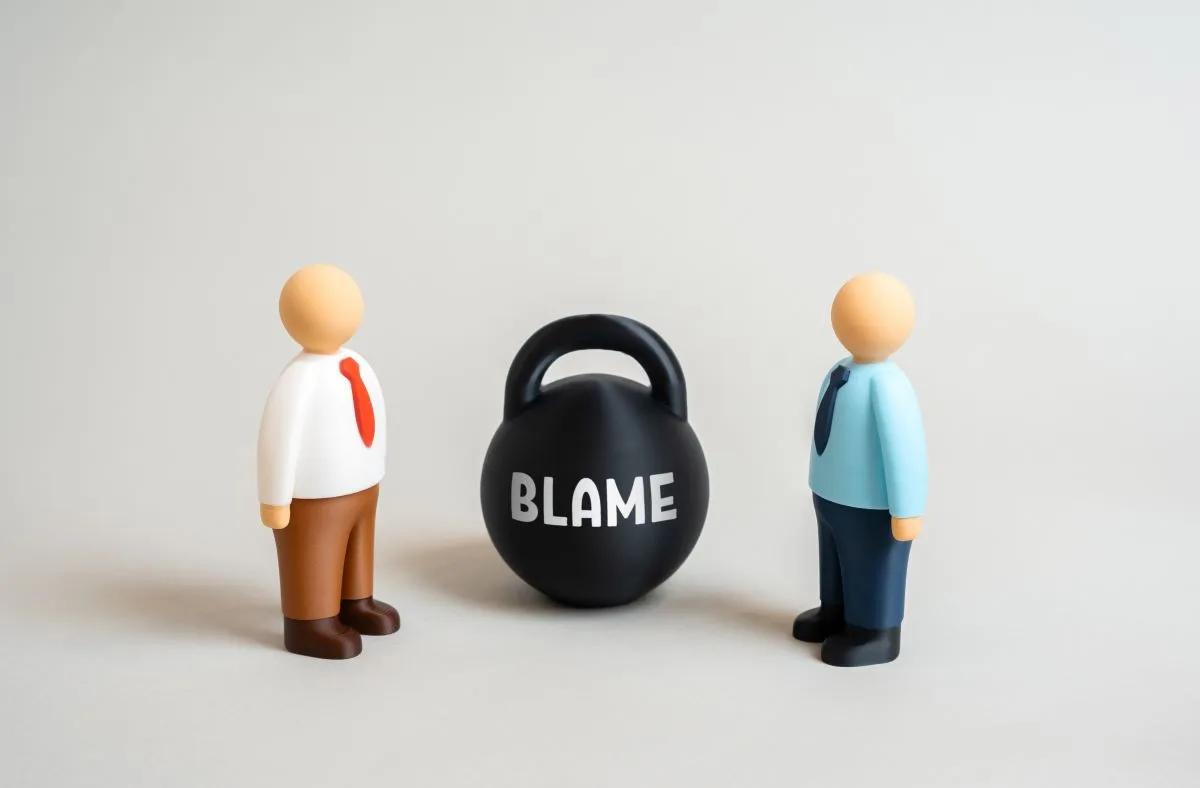 Toy people stand next to kettlebell labeled “Blame” as they argue over fault.