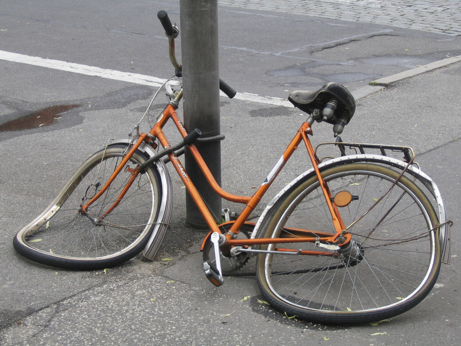 A wrecked orange bicycle locked to a street pole.