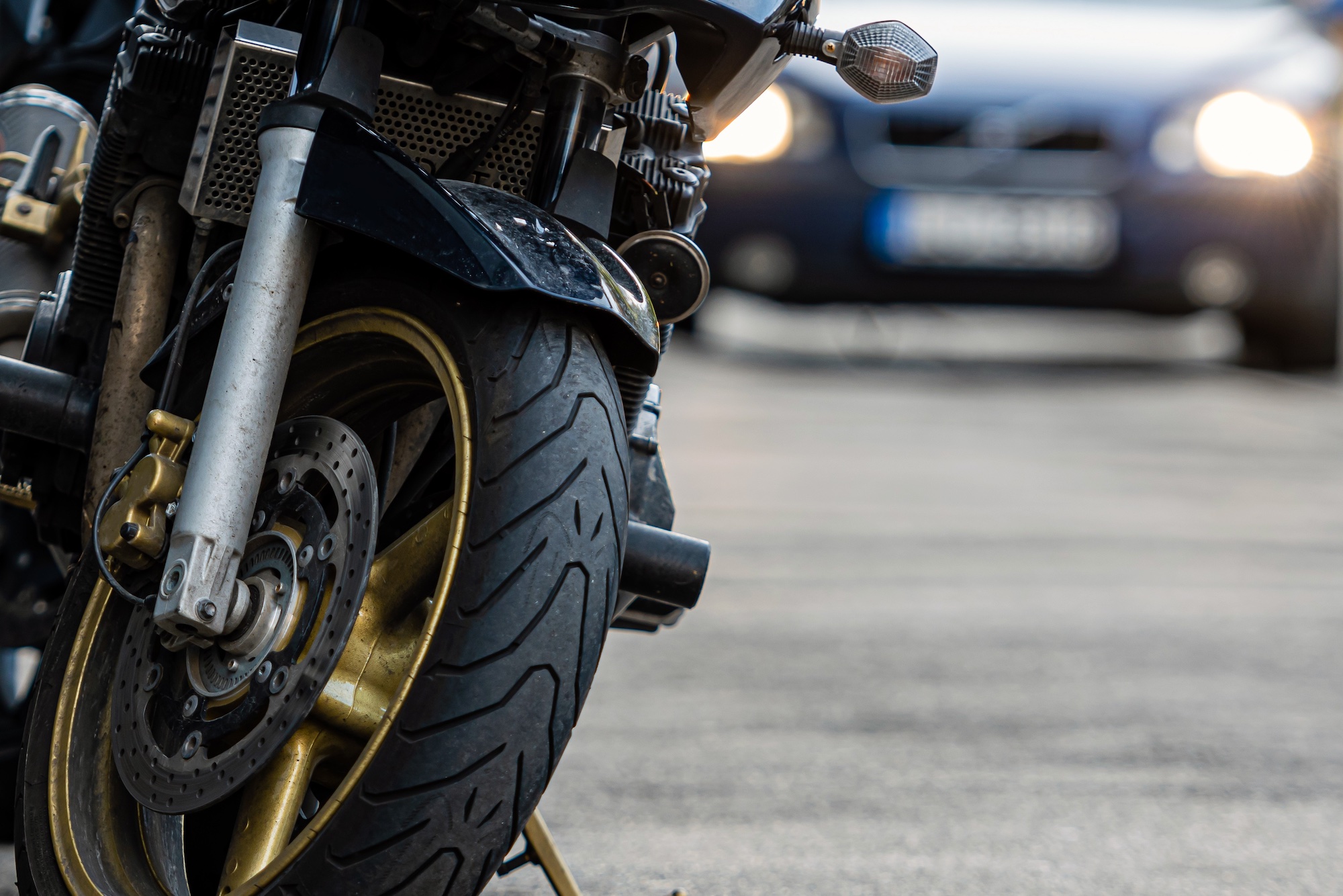 On the left, the front of a motorcycle is seen on the side of the road. To the right, an out-of-focus car with its headlights on.