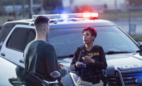 A policewoman taking notes for a police report, while talking to a young man after a car accident.