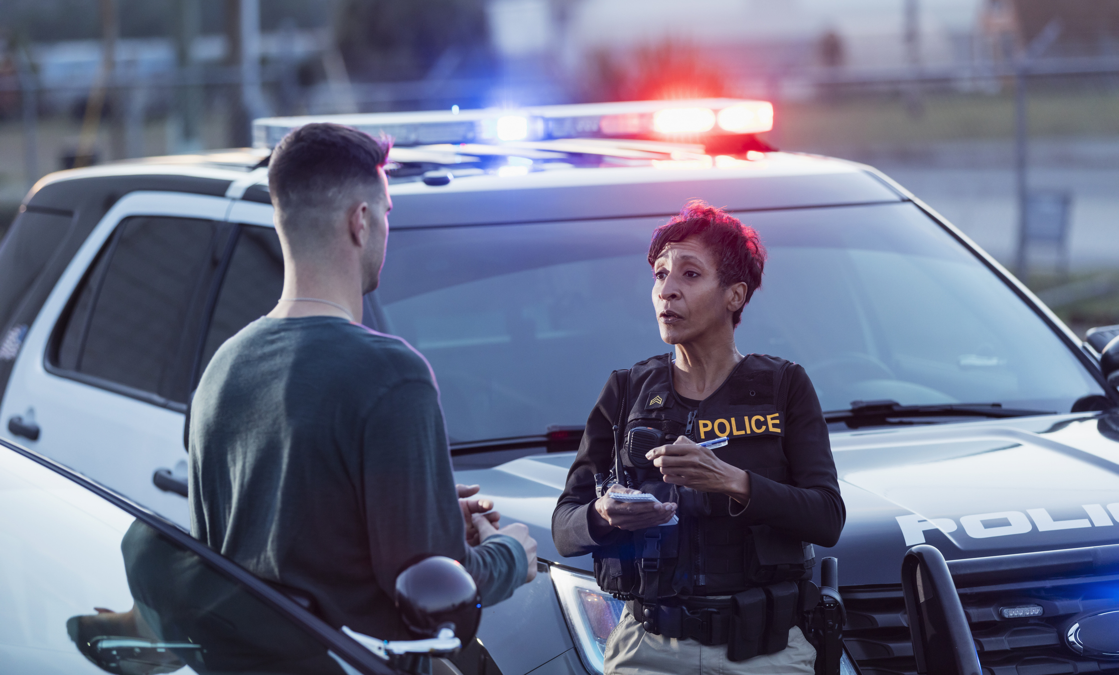 A policewoman taking notes for a police report, while talking to a young man after a car accident.