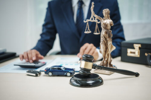 A lawyer at their desk, typing on a calculator while reviewing paperwork. A car key fob, a blue model car, a gavel, a Lady Justice Statue, and a briefcase are also sitting on the desk.