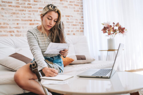 A woman with an amputated leg takes notes as she reads her bills. Her laptop sits open on the table in front of her.