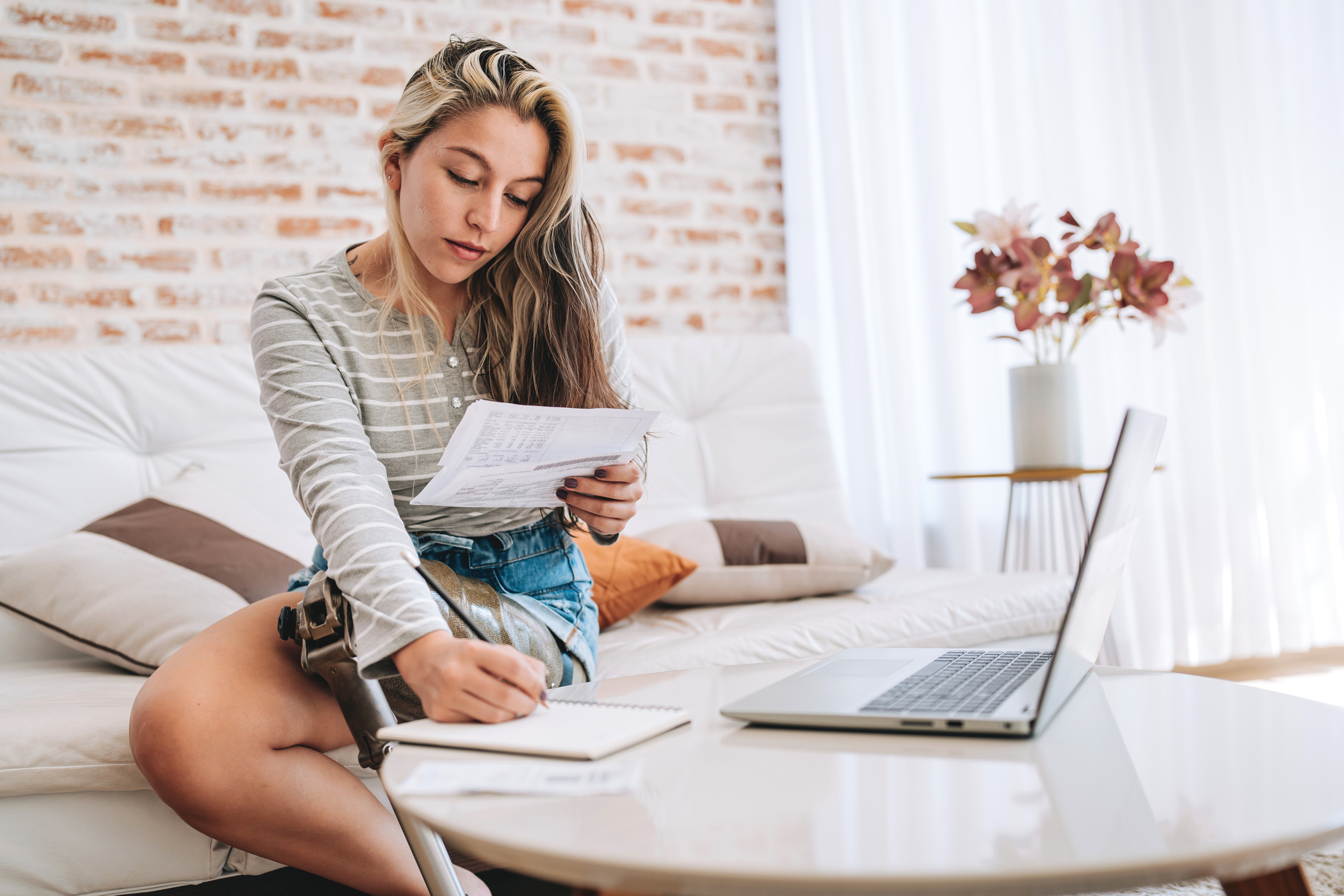 A woman with an amputated leg takes notes as she reads her bills. Her laptop sits open on the table in front of her.